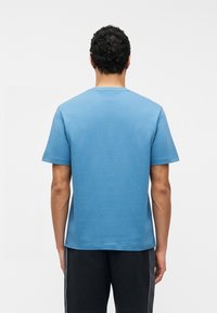 Homme aux cheveux bouclés foncés, debout de dos, portant un t-shirt bleu uni à manches courtes et un short noir avec des coutures latérales blanches.