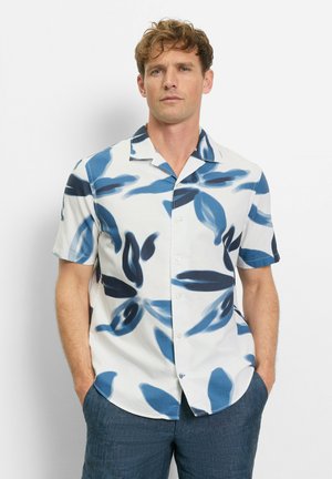 Mann mit kurzen lockigen Haaren, der ein weißes Kurzarmhemd mit blauen Blättermustern und blaue Hose trägt, Hände in den Taschen.