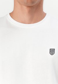 Weißes Baumwoll-T-Shirt mit rundem Halsausschnitt, versehen mit einem kleinen schwarzen gestickten Wappenlogo auf der linken Brust. Weicher Stoff, schlichtes Design.