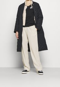 Osoba oblečená v černém prošívaném dlouhém kabátu, béžových kalhotách, černém tričku Nike, vrstvené košili a černobílých teniskách, stojí se založenýma rukama v kapsách.