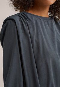 Haut gris foncé avec des manches longues drapées et une texture douce, agrémenté de plis subtils aux épaules et d'un col rond.