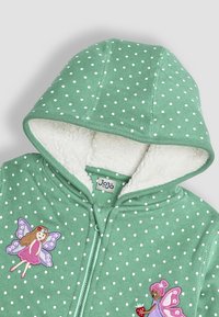 Zelená mikina s kapucňou s bielymi bodkami, s bielou fleecovou podšívkou, ktorá má na prednej strane dve vyšívané designy víl a zapínanie na zips.