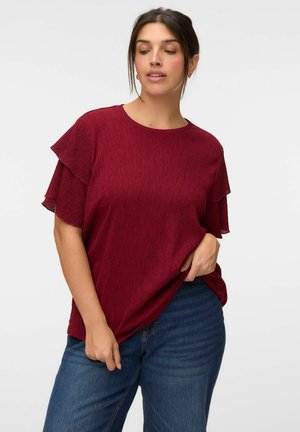 Femme portant un chemisier rouge texturé à manches courtes avec des manches à volants superposés et un jean bleu, posant avec une main dans la poche.