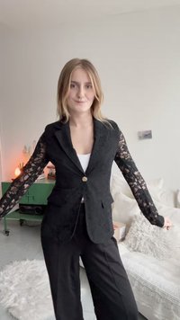 Čierny blazer s čipkovými rukávmi, jedným zlatým gombíkom a textúrovanou tkaninou. V páre s čiernymi nohavicami, vystavený v neformálnom prostredí.