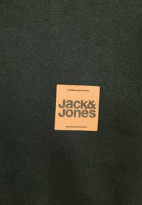 Dark green cotton fabric featuring a square tan label with "Jack & Jones" printed in bold, dark green text. Smooth texture, casual design.