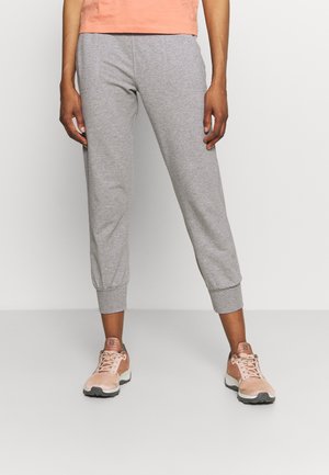 Persoon draagt lichtgrijze joggingbroek met omgeslagen enkels en perzikkleurige sportschoenen, staand tegen een effen lichte achtergrond.