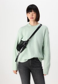 Pull en maille côtelée vert menthe à manches longues, associé à un jean gris foncé. Un petit sac bandoulière noir avec des détails de boutons est porté en travers du corps.