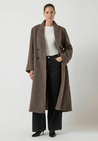 Abrigo largo marrón con un material texturizado similar a la lana, diseño de doble botonadura, botones grandes, combinado con pantalones negros de pierna ancha y cintura alta.