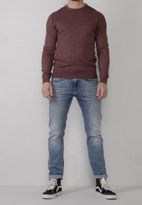 Uomo che indossa un maglione bordeaux, jeans azzurri chiari arrotolati ai polsi e sneaker alte nere con lacci bianchi, in piedi contro un muro semplice.