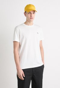 Camiseta blanca de algodón con mangas cortas y cuello redondo. Combinada con una gorra de béisbol amarilla. Diseño simple, sin patrones visibles.