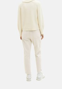 Personne portant un pull crème, un pantalon beige clair, des chaussettes blanches et des baskets blanches épaisses, vue de dos sur un fond uni.