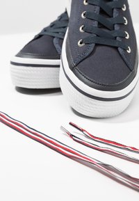 Gros plan de baskets en toile bleu marine avec semelles en caoutchouc blanches et lacets rayés noirs, rouges et blancs posés à côté.