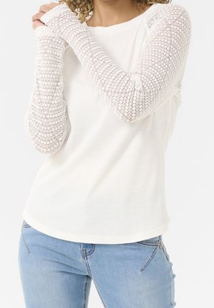 Vrouw die een wit top met lange mouwen en kanten mouwen draagt en een lichtblauwe spijkerbroek, met de armen over de borst gekruist, tegen een effen achtergrond.