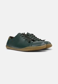 Zapatillas de cuero verde con textura suave, punta redonda y suela de goma marrón en contraste. Presenta cordones negros y un detalle de costura simple.