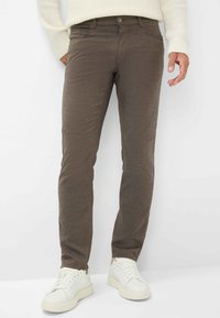 Braune, schmal geschnittene Hose aus strukturiertem Stoff mit einem dezenten Muster. Verfügt über Vordertaschen und einen Knopfverschluss. Kombiniert mit weißen Sneakern.