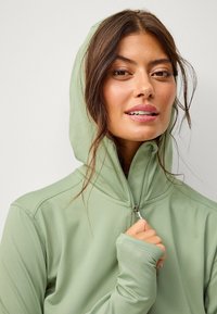 Kapuce s zipem v světle zelené barvě, s jemnou texturou, přiléhavým střihem a otvory na palce na manžetách. Zip je viditelný vpředu.