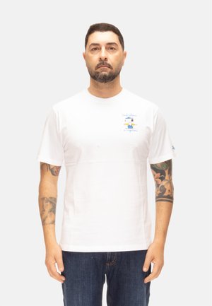 Uomo con capelli corti e scuri e barba in piedi, indossa una maglietta bianca con un piccolo disegno colorato e jeans blu scuro, braccia tatuate.