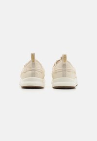 Jasno beżowe sneakersy z gładką tekstylną cholewką, wyściełanym kołnierzem oraz kontrastującą białą podeszwą z teksturowanym wzorem gripu. Złote logo marki z tyłu.