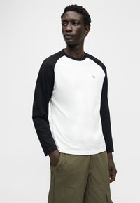 Λευκό βαμβακερό μακρυμάνικο μπλουζάκι με μαύρα raglan μανίκια, με μικρό λογότυπο στο στήθος και άνετη εφαρμογή, σε συνδυασμό με χακί παντελόνι cargo.