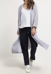 Cardigan aperto lilla chiaro con maniche trasparenti, abbinato a una maglietta bianca con scollo a V e pantaloni blu navy. Il modello indossa sneakers bianche.