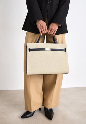 Persoon in beige wijde broek en zwarte blazer houdt rechthoekige beige handtas met zwarte handvatten en afwerking vast, staande op een lichte vloer.