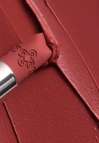 Matte läppstift i varm mauve med präglad logotyp och metallisk silverbas. Krämig textur visad mot en matchande utstrykning i bakgrunden.