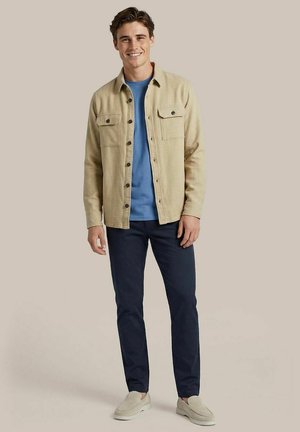 Lächelnder Mann, der ein beiges Hemd mit Knöpfen über einem blauen T-Shirt, marineblaue Hosen und beige Slipper trägt und vor einem einfarbigen Hintergrund steht.