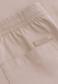 Pantaloni elasticizzati beige con vita elastica arricciata e tasca posteriore. Presentano un piccolo tag in rilievo con il logo di Calvin Klein.