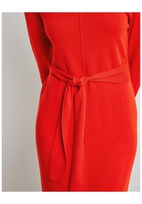 Robe en maille rouge avec une ceinture nouée, présentant une texture lisse et un design ajusté. Encolure ronde et manches longues visibles.