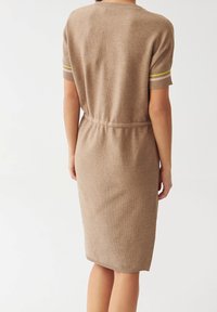 Vue de dos d'une personne portant une robe tricotée beige jusqu'aux genoux avec des manches courtes et un détail de ceinture, se tenant devant un fond blanc.