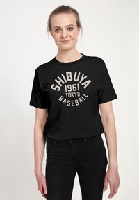 Žena oblečená v černém tričku s nápisem "SHIBUYA 1961 TOKYO BASEBALL" potištěném béžovou barvou, v kombinaci s černými džíny, stojí na bílém pozadí.