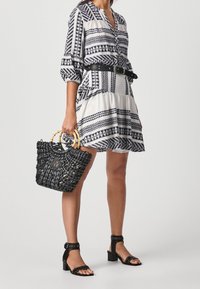 Donna che indossa un vestito con un motivo geometrico bianco e nero, cintura nera, sandali neri con tacco alto, e tiene una borsa nera intrecciata con manici in bambù.