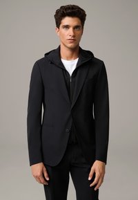 Czarna marynarka nałożona na czarną bluzę z kapturem na zamek, o klasycznym kroju, gładkiej strukturze i subtelnym wykończeniu kieszeni.