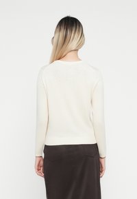 Cream-colored sweater with long sleeves, ribbed hem, and subtle embellishments on the back. Worn over a dark, tailored skirt.