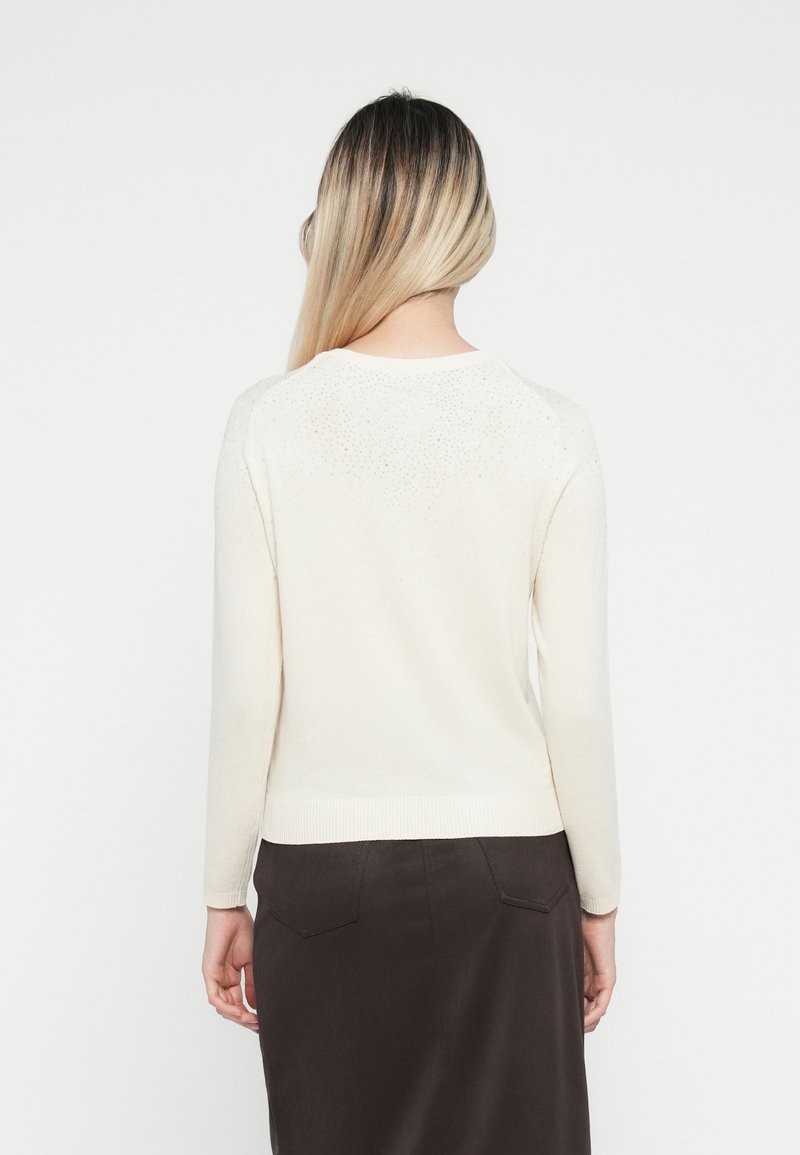 Jersey de color crema con mangas largas, dobladillo acanalado y sutiles adornos en la parte posterior. Lleva encima de una falda oscura, entallada.