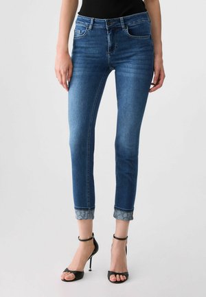 Jeans blu slim fit con risvolti, indossati con sandali neri con cinturino alla caviglia e tacco alto, mostrati dalla vita ai piedi su uno sfondo chiaro.