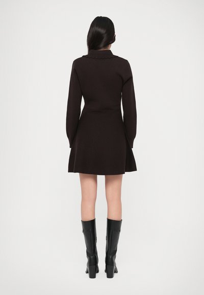 Robe en tricot noir avec un col festonné, des manches longues et un ourlet évasé. Portée avec des bottes noires à talons hauts, mettant en avant une texture lisse et un design structuré.