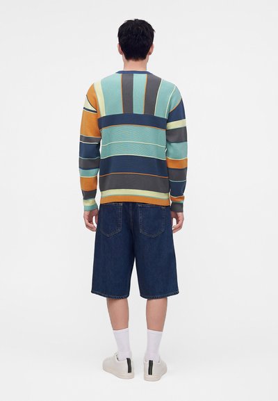 Homme portant un pull rayé multicolore, un short en jean bleu foncé jusqu'aux genoux, des chaussettes blanches et des baskets blanches, debout de dos.