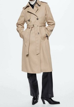 Beige kaksirivinen trenssitakki vyöllä mustujen nahkahousujen ja mustien korollisten saappaiden päällä, kädet taskuissa, valkoisella taustalla.