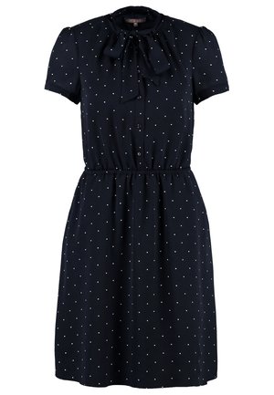 Femme marchant avec une robe à pois navy et un nœud, un blazer beige, des collants noirs, des talons noirs et portant un sac à main noir.