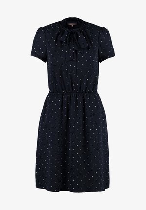 Kvinde går i en navyblå kjole med prikker og sløjfe, beige blazer, sorte strømpebukser, sorte heels og bærer en sort håndtaske.