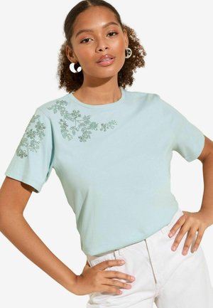 Camiseta de algodón azul claro con mangas cortas, que presenta bordado floral brillante en un hombro. Textura suave y corte clásico.