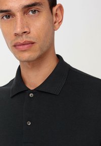 Zwarte poloshirt met een gestructureerde stof, twee knopen bij de kraag en een klassiek ontwerp. Close-up weergave toont de details van de kraag.