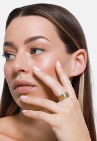 Anello d'oro con texture rigata indossato su un dito. Il modello presenta un tono di pelle soft, trucco neutro e smalto per unghie rosa chiaro su uno sfondo bianco.