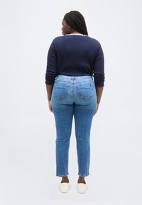 Γυναίκα με πλεξουδάκια στα μαλλιά, που φοράει μπλε μπλούζα με μακριά μανίκια, μπλε τζιν και λευκά παπούτσια χωρίς κορδόνια, στέκεται με την πλάτη σε λευκό φόντο.