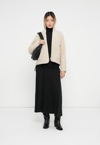 Μπεζ φλις ζακέτα με μεγάλες τσέπες, φορεμένη πάνω από μαύρο πουλόβερ και μακριά μαύρη φούστα. Μαύρες μποτες με τακούνι ολοκληρώνουν το σύνολο.