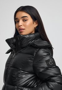 Czarna kurtka puchowa z wysokim kołnierzem, teksturowanym materiałem, zapięciem na zamek i logotypem na lewym rękawie.