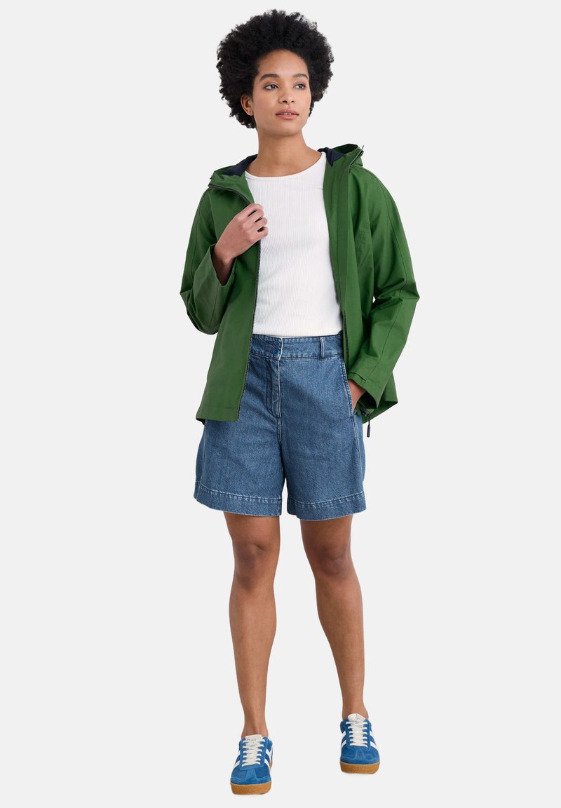 Zielona wodoodporna kurtka, biała bluzka z prążkowanej tkaniny, niebieskie szorty dżinsowe i niebieskie sneakersy z białymi sznurówkami. Swobodny strój odpowiedni do noszenia na świeżym powietrzu.