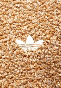 Καφέ υφασμάτινη υφή με λευκό κεντητό λογότυπο που απεικονίζει τρία σχήματα φύλλων και δύο οριζόντιες γραμμές από πάνω του.