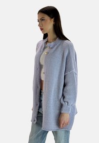 Cárdigan de punto en lavanda claro con cuello redondo, dos botones y mangas sueltas. Lleva puesto sobre una camiseta cropped blanca y unos jeans de color azul claro.